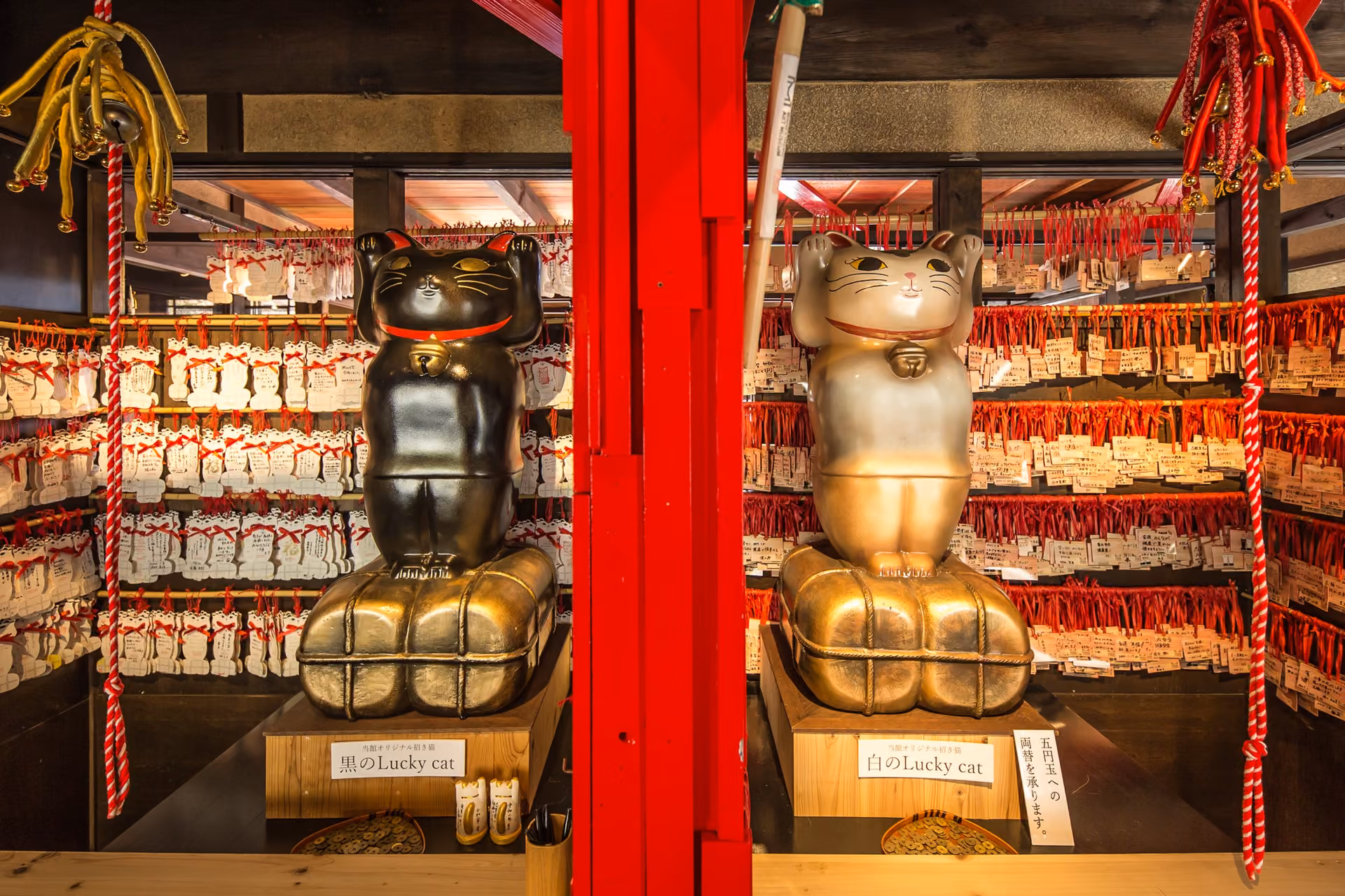 A black and a white Lucky Cat statue with raised paws, sitting on gold rice bales in front of rows of wish plaques hung on red cords