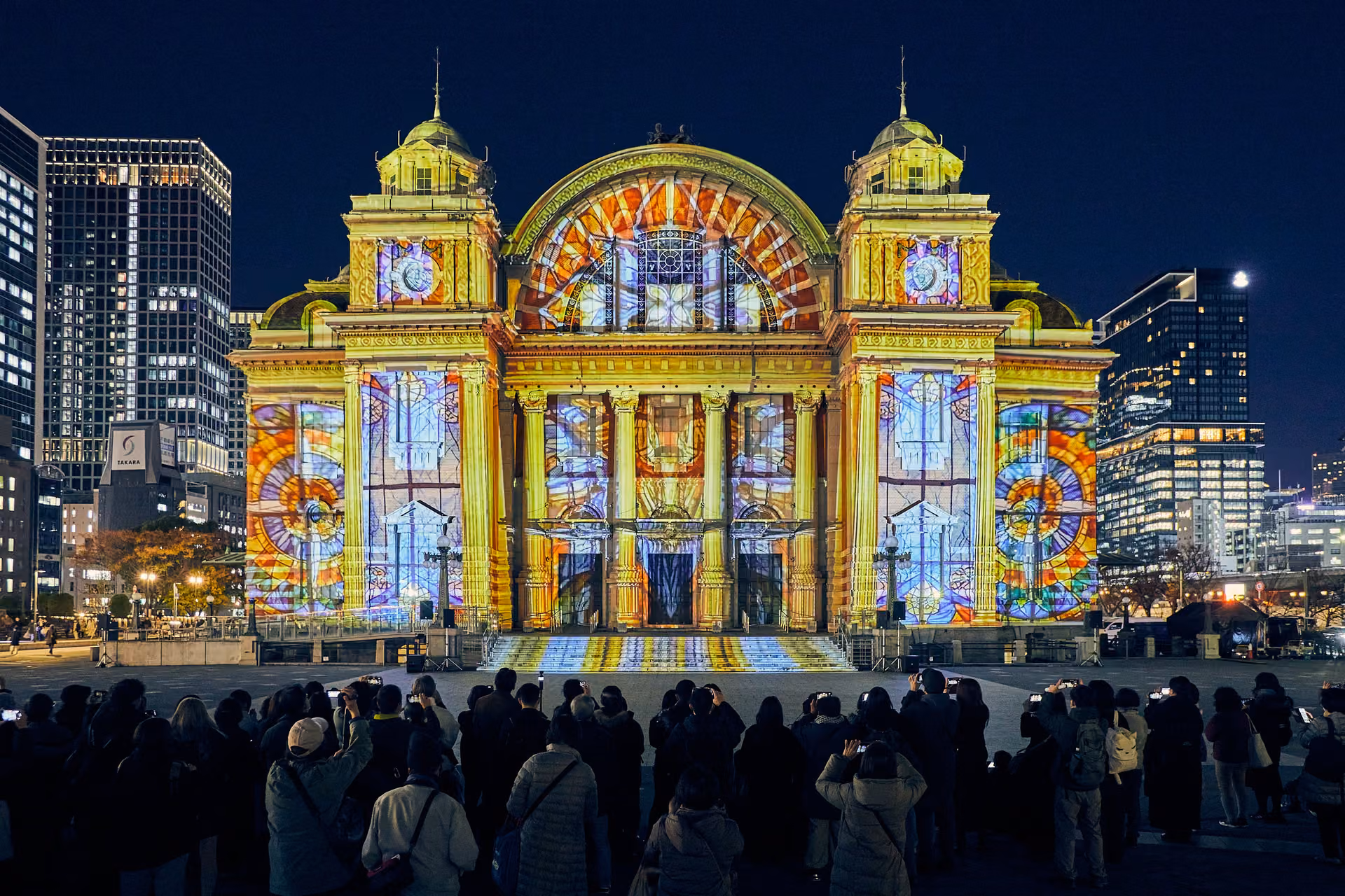 The red-brick front of Osaka City Central Public Hall at night, covered in projected yellow and blue panels, a crowd watching