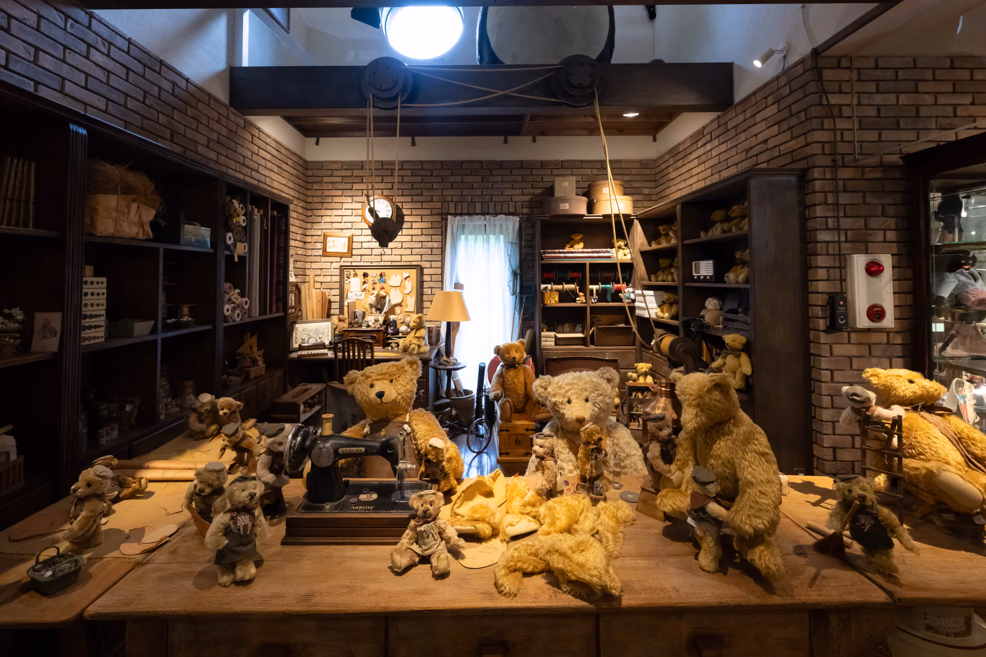 Old golden teddy bears gathered around a sewing machine in a display set up like a bear maker's workshop, with brick walls and wooden shelves