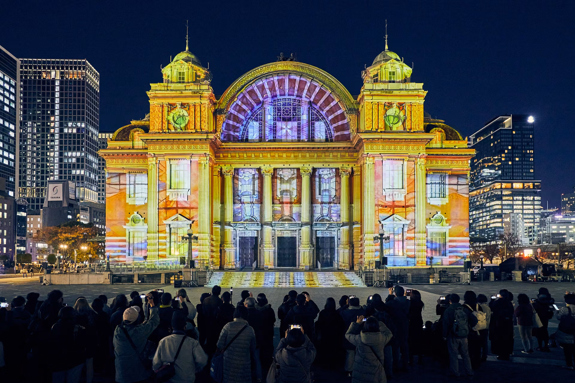 The front of Osaka City Central Public Hall lit gold, with a purple glow on the central arch and viewers in the foreground
