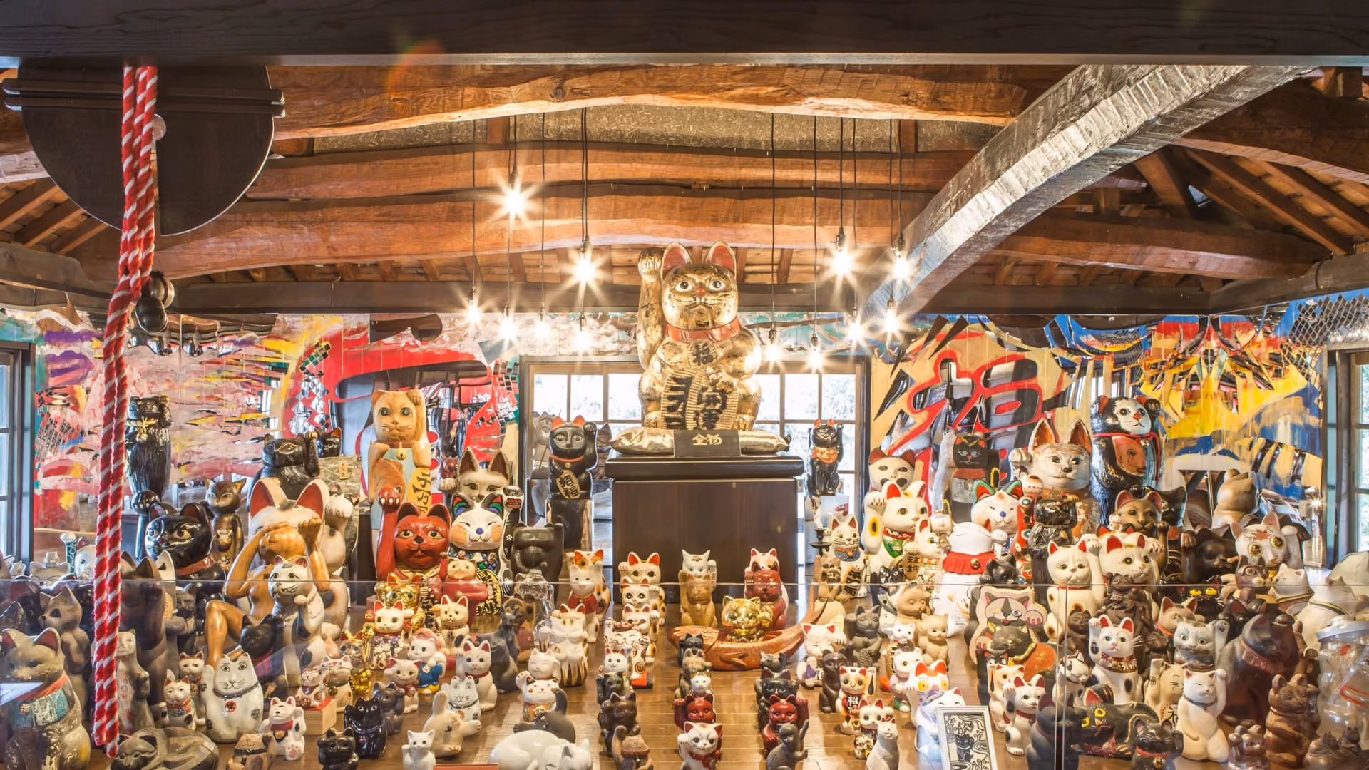 Maneki neko of many sizes and colors on display under the wooden roof beams of the Manekineko Art Museum, with a large gold cat at the center