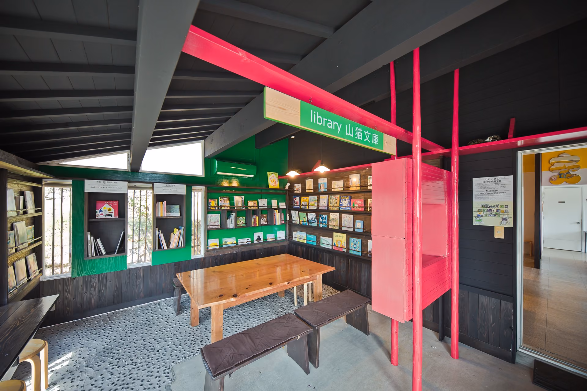 The library Yamaneko Bunko, with a wooden table and benches, green and black bookshelves and a pink frame under a green sign