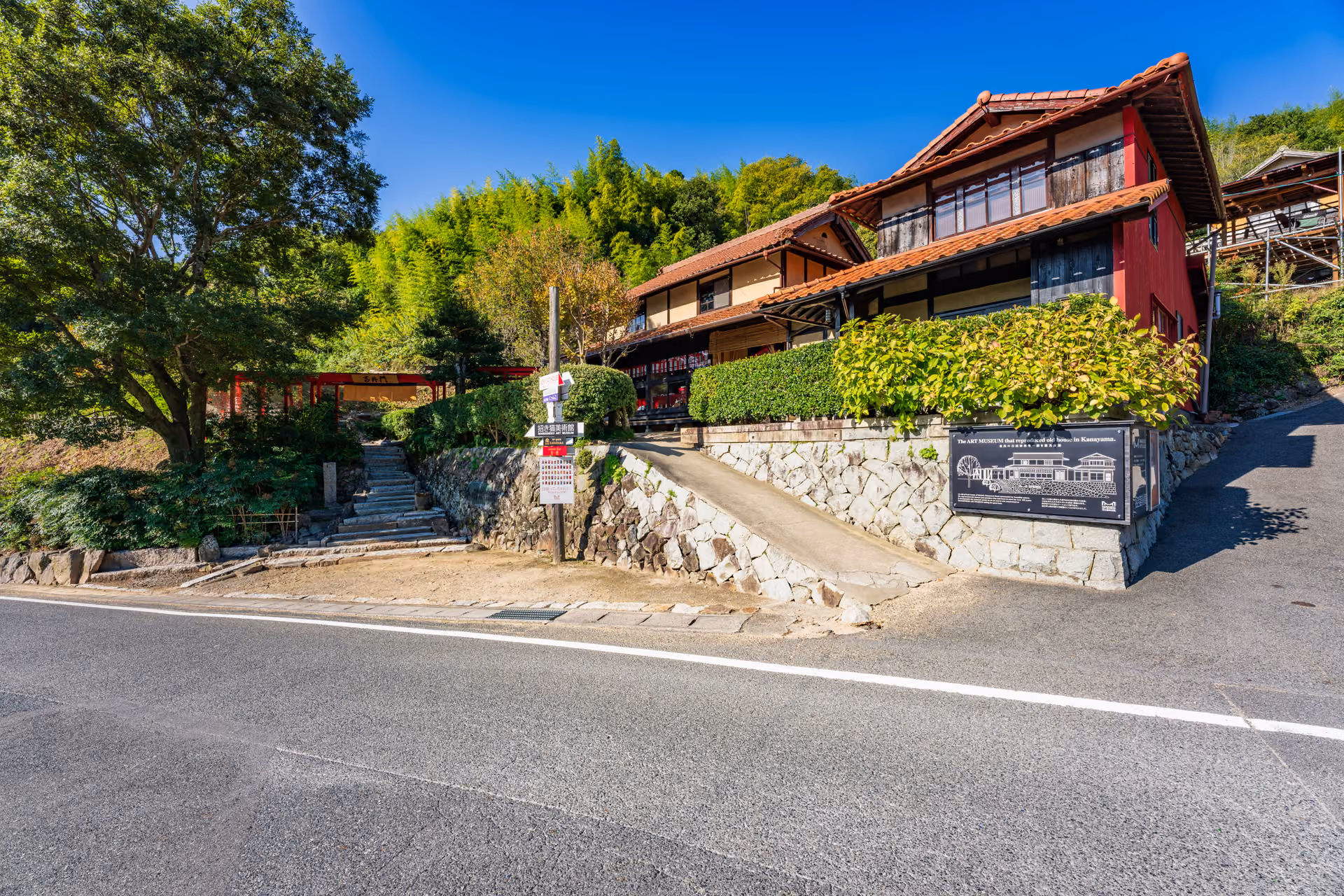 The two-storey main building of the Manekineko Art Museum, with red walls and a tiled roof, above a stone wall by the road